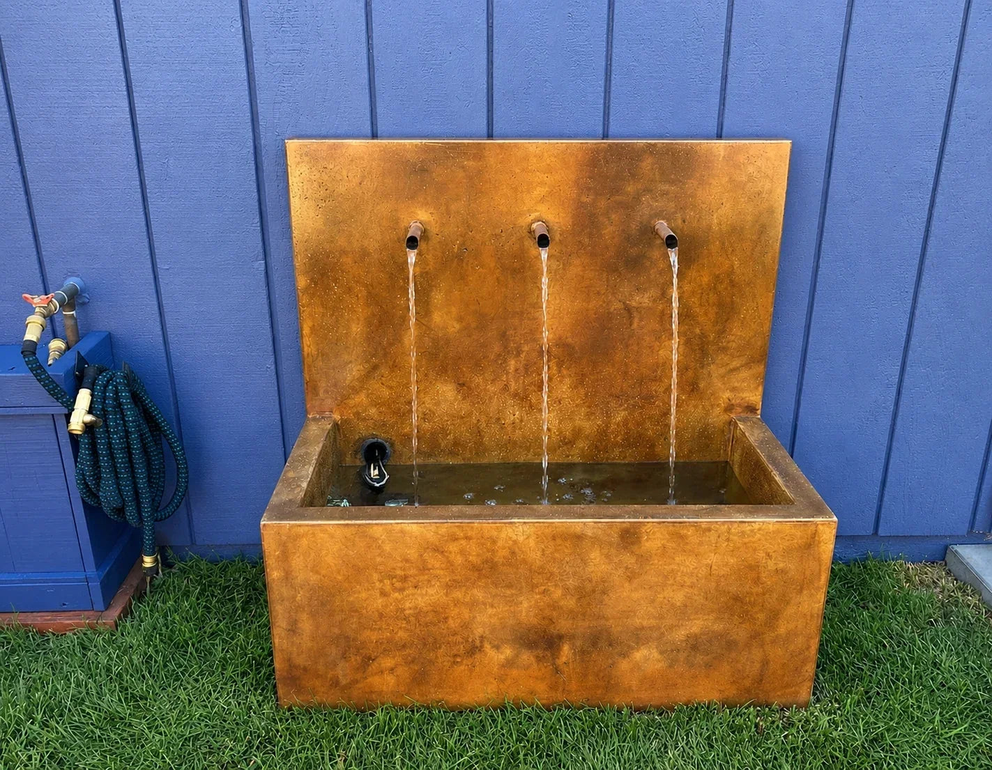 A three-spout concrete wall fountain colored in rust and amber tones, water running into its basin against a blue wall