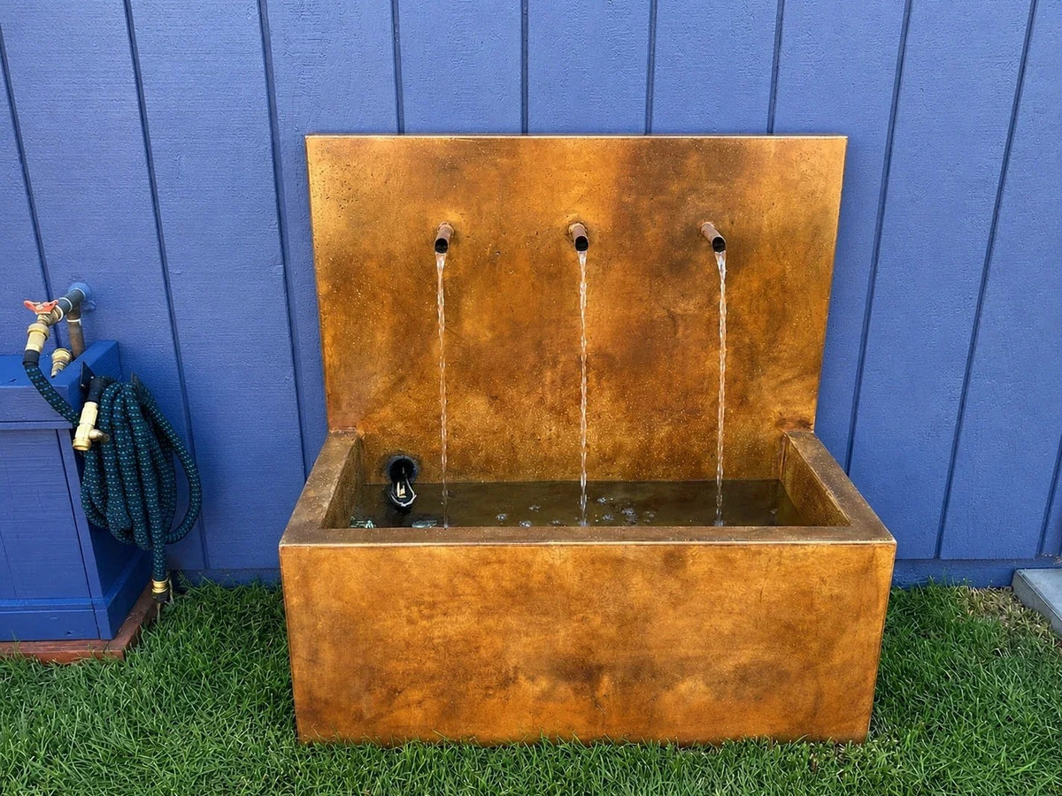 A three-spout concrete wall fountain colored in rust and amber tones, water running into its basin against a blue wall