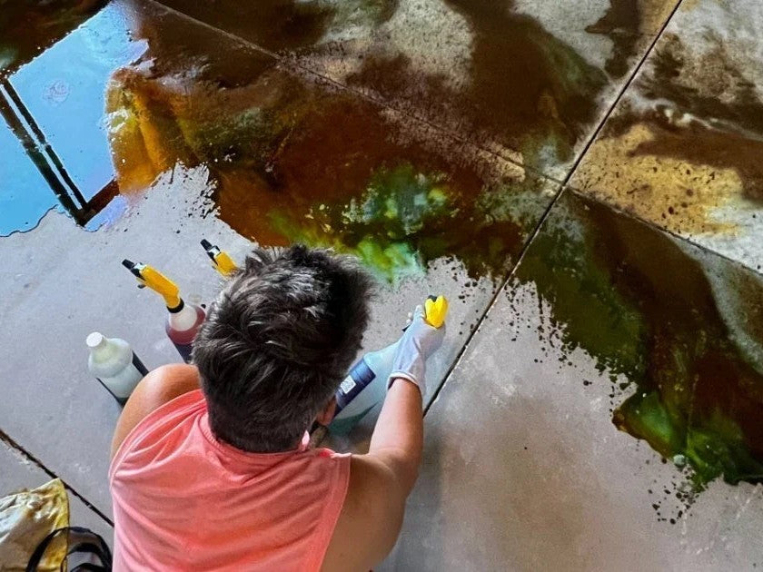 A DIYer spray-applying acid stain wet-on-wet to a concrete floor, green and brown color pooling and blending across the slab
