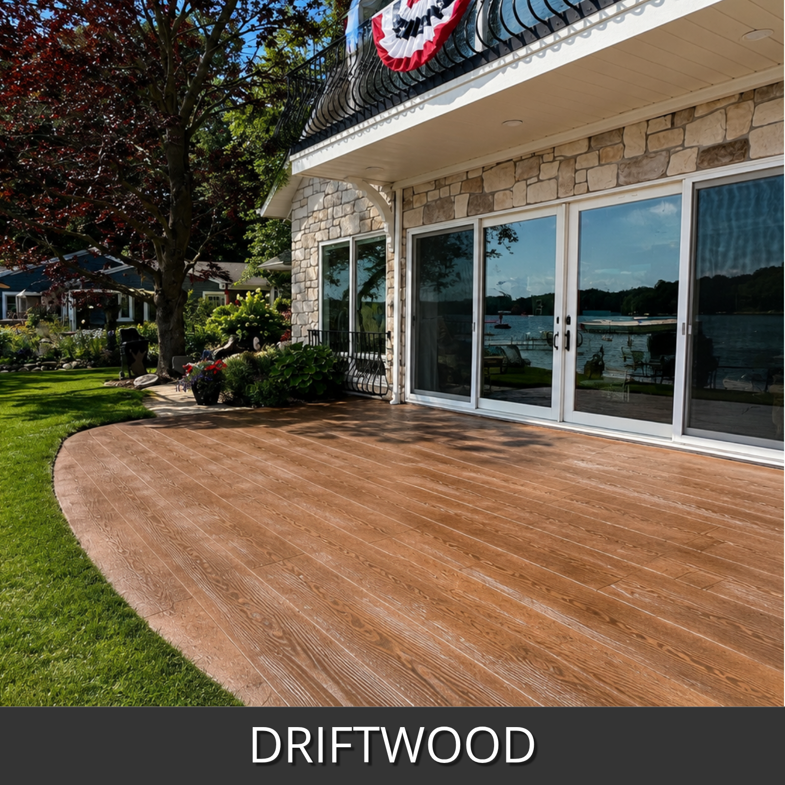 A curved outdoor deck made of driftwood faux wood stamped concrete planks installed in front of a stone house with large glass doors, with grass, trees, and a lake visible in the background