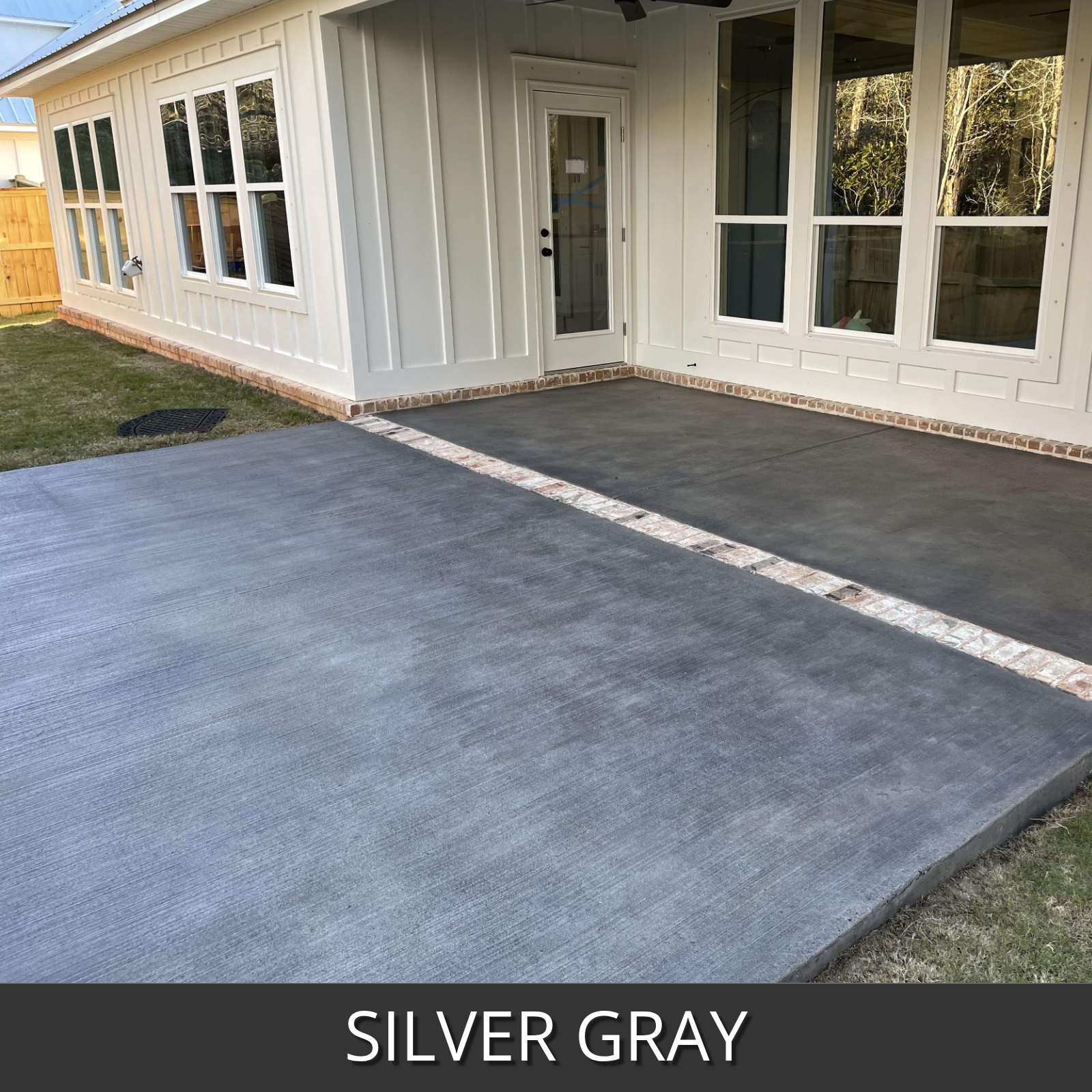 Broom finished concrete patio in front of a house, shown in a silver gray finish. The patio is bordered by a decorative stone strip and is set next to a white-sided house with large windows.