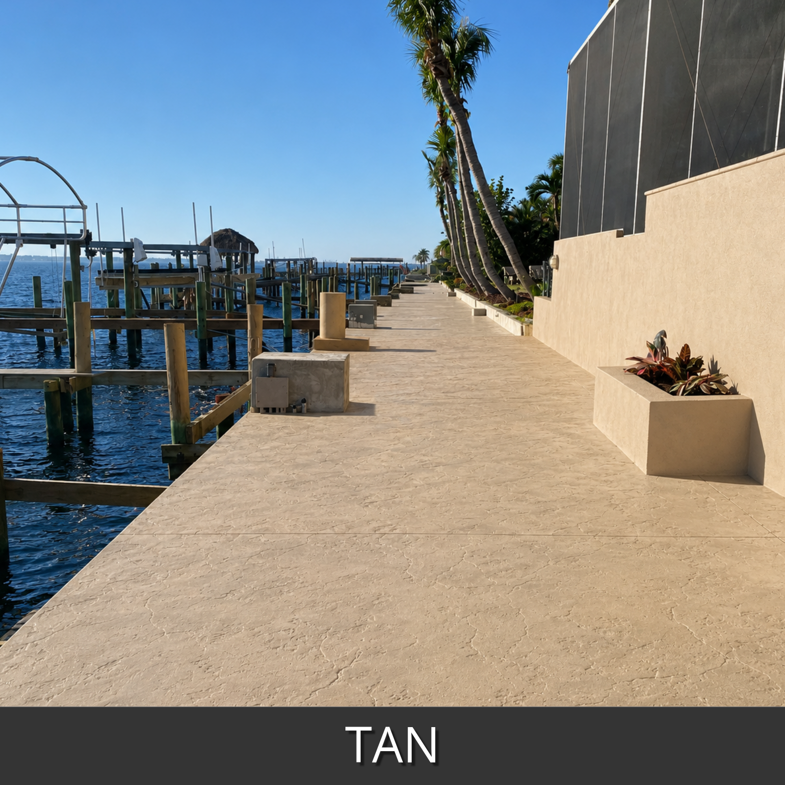 A tan colored textured concrete deck running along a waterfront promenade, with blue water, wooden docks, palm trees, a beige building wall
