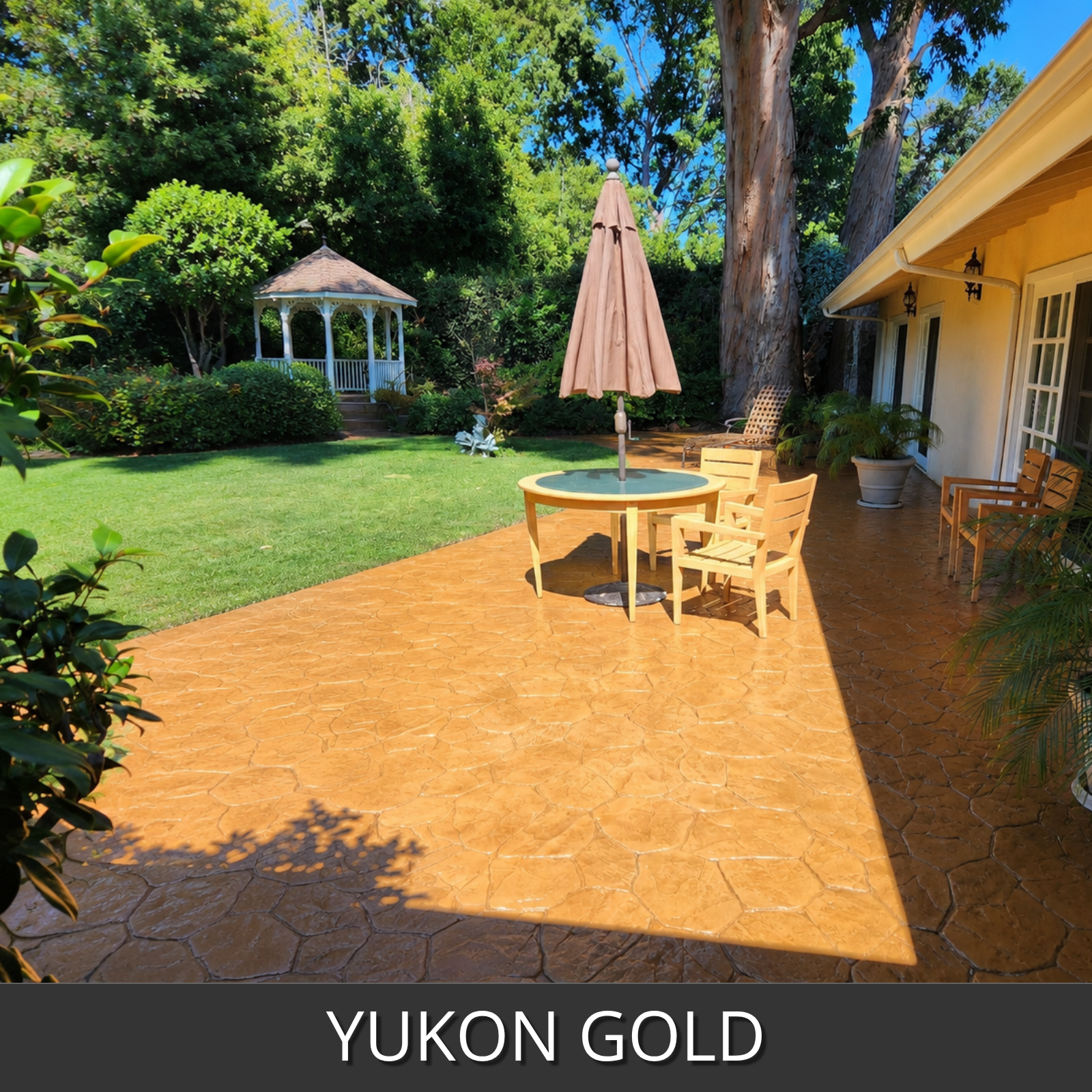 A stamped concrete patio in a backyard setting, featuring a textured stone-like pattern with a yukon gold color The patio is furnished with a round table, wooden chairs, and a closed patio umbrella, with a green lawn, trees, and house visible in the background.