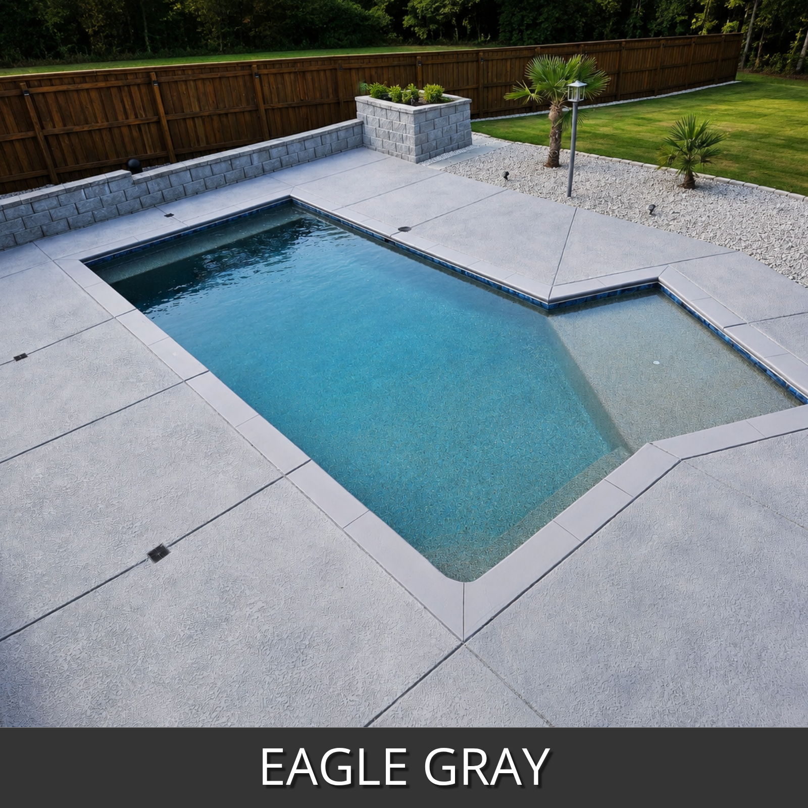A backyard pool with eagle gray concrete decking and a gray stone wall, shown in a landscaped yard with grass, fence, and small plants.