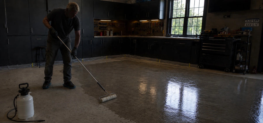 Contractor rolling concrete sealer onto a stained garage floor using a 3/8 inch nap roller and extension pole, working in overlapping W-shaped passes from the back wall toward the door