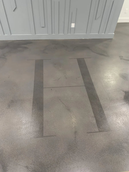 A concrete floor with a large, stylized letter "H" stenciled onto the surface, set against a faux polished finish with subtle color variations
