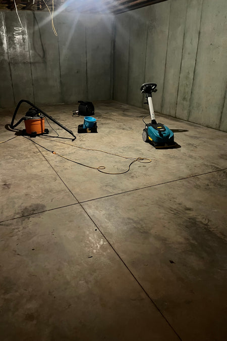 A basement with unfinished concrete floors, cleaning tools, and a scrubber preparing the surface for Malayan Buff and Seagrass acid stain