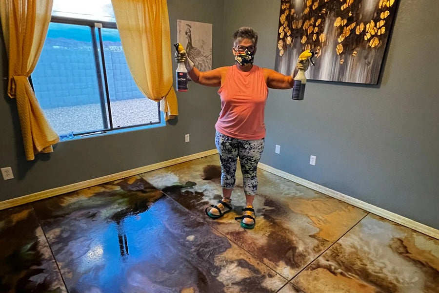A DIYer in a mask holds two pump sprayers over a freshly acid-stained concrete floor, the wet surface mottled in amber, brown and dark tones