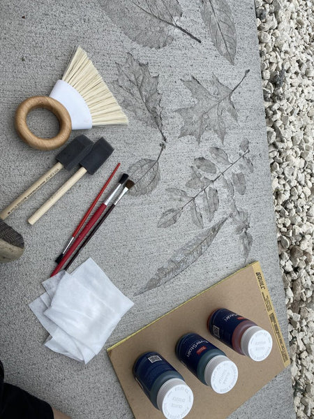 Close-up of tools including brushes, foam applicators, and EverStain acid stain bottles, with leaf stencils on concrete in the background