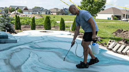 A person spraying an acid stain solution on a concrete surface outdoors, with swirling blue-green patterns forming on the ground