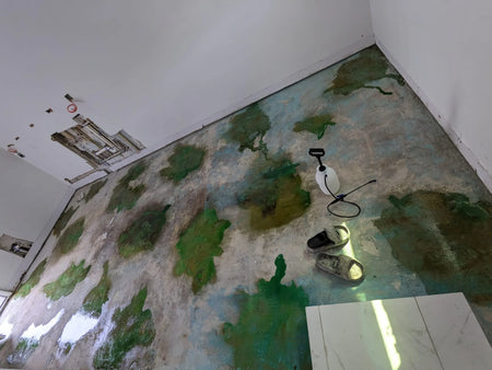 A concrete floor with large splotches of green stain applied, with a sprayer and footwear visible on the floor