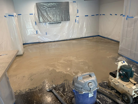A room with plastic sheeting covering the walls, showing a concrete floor being ground down with a floor grinder, with debris scattered around