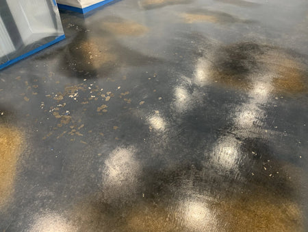 A concrete floor with patches where stain and wax have flaked off, exposing bare concrete. The surface is smooth and reflective, showing poor stain adhesion