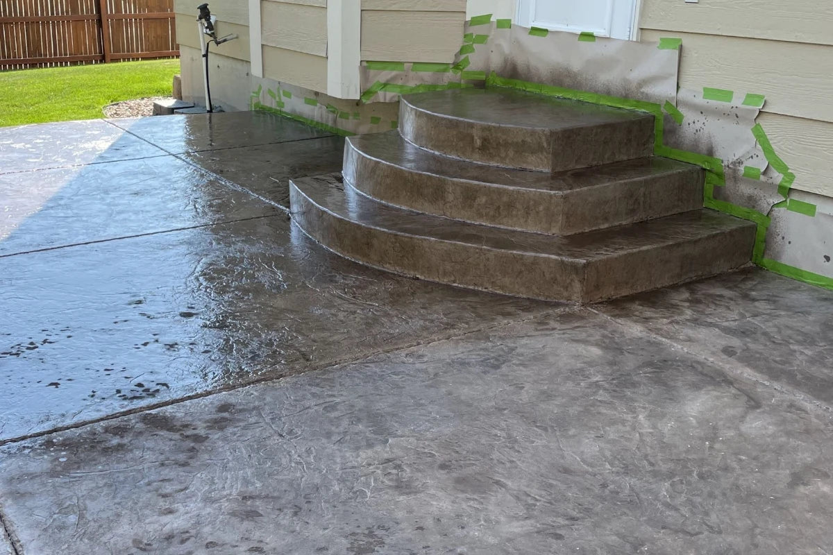 A curved stamped concrete patio being stained with Driftwood Antiquing stain, bordered by grass and a wooden fence