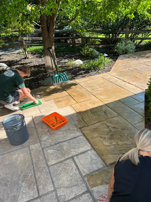 Two people applying warm Tweed color to a stamped concrete patio with a foam applicator, tray, and bucket beside an untreated gray section