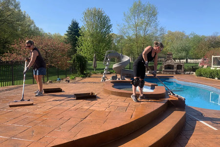 Two homeowners rolling a second coat of Yukon Gold EasyTint onto a stamped concrete pool deck beside a spa
