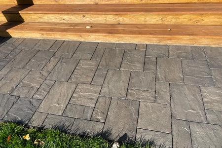 Finished charcoal stamped concrete pavers running beside broad wooden deck steps, grass, and a protected porch post