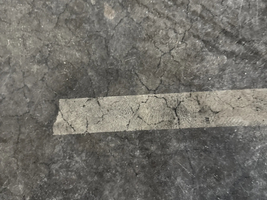 Painter's tape lifted a clean strip of sealer and color from a dark stained concrete floor, leaving bare gray concrete where the tape sat