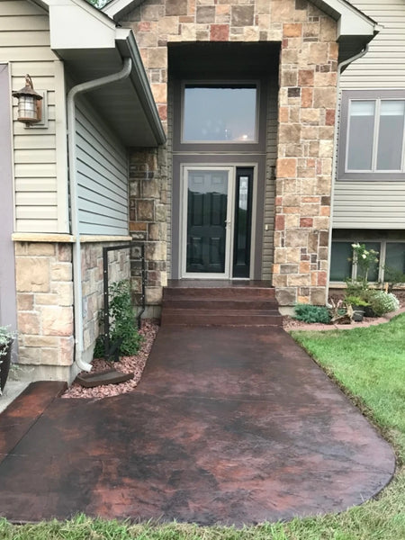 Finished dark rusty-brown and charcoal stamped concrete walkway curving through grass toward the front steps of a stone-trimmed house