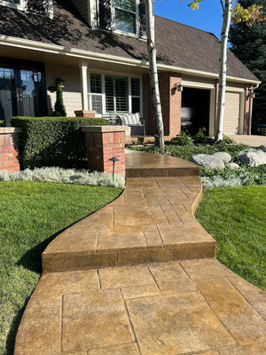 Finished warm Tweed-colored stamped concrete walkway and steps curving through a landscaped front yard toward a house