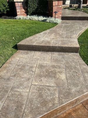 Close view of a stamped concrete walkway during refinishing, showing faded gray-brown color, stamped joints, and darkened step risers