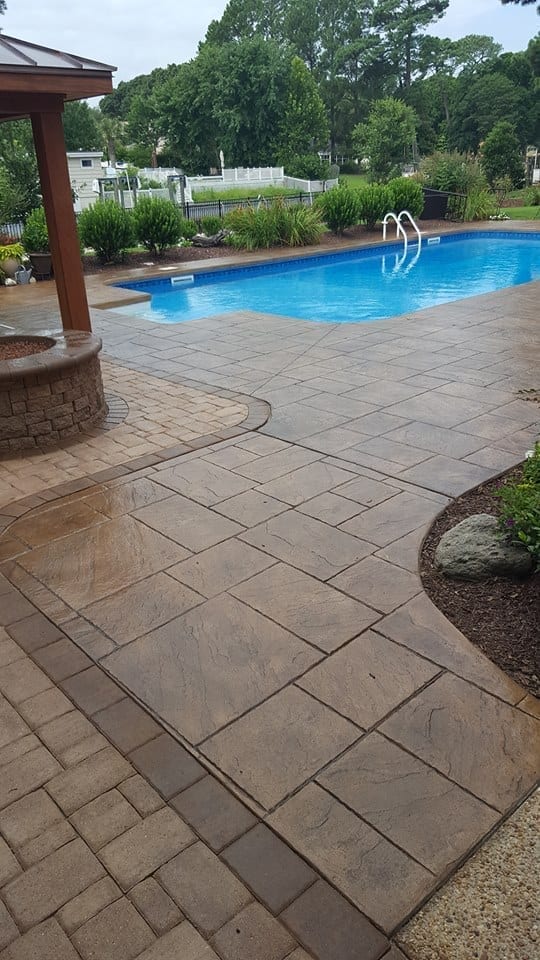 A stunningly detailed stamped concrete pool deck displaying a rich blend of Aztec Brown, Charcoal, and Cafe Royale antiquing stains, replicating the look of natural stone.