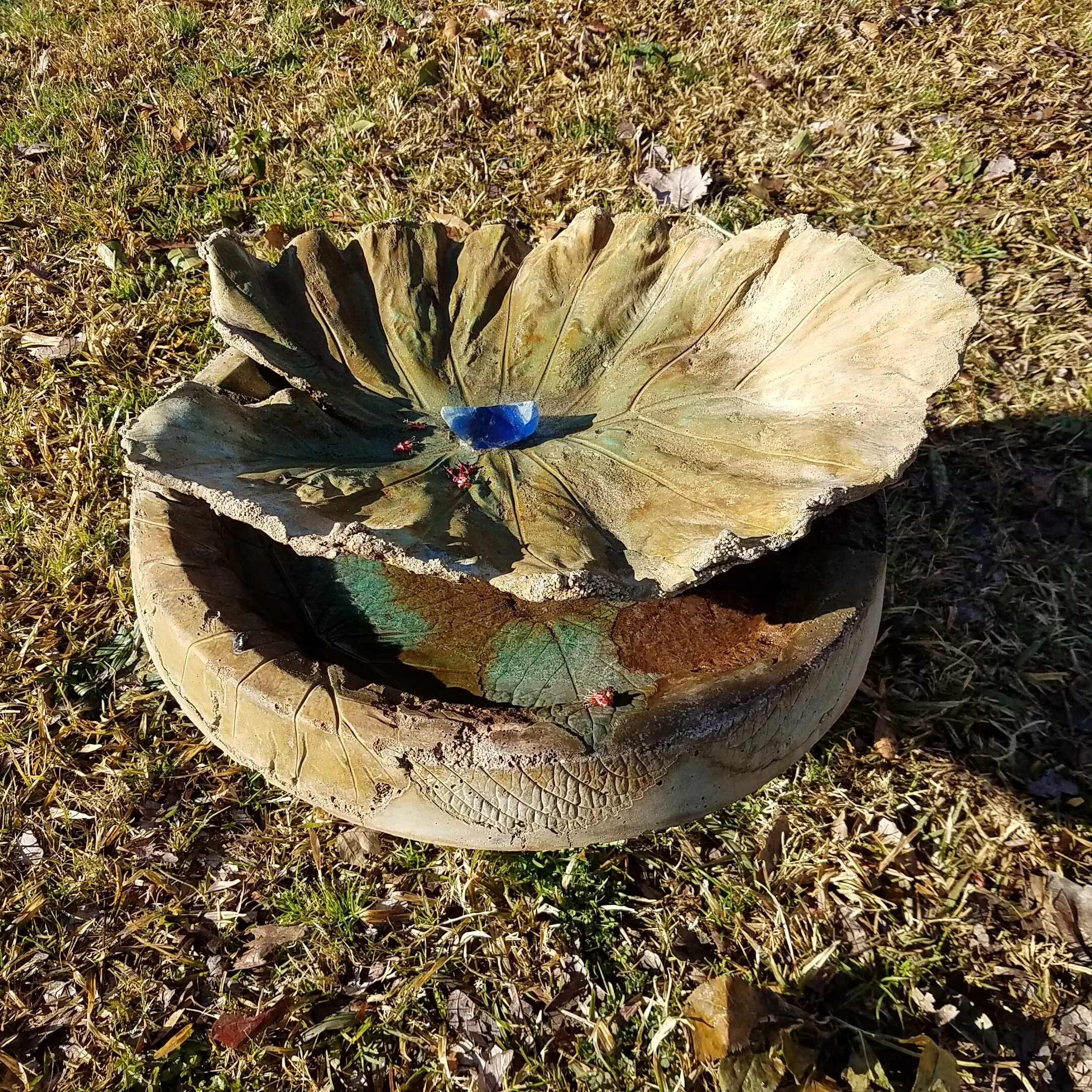 A picturesque bird bath stained with Shifting Sand acid stains for the base and Azure Blue accents highlighting its artistic features. The bird bath is structured like a bowl, with a large, intricately designed concrete leaf mounted on top, providing an inviting spot for the birds.