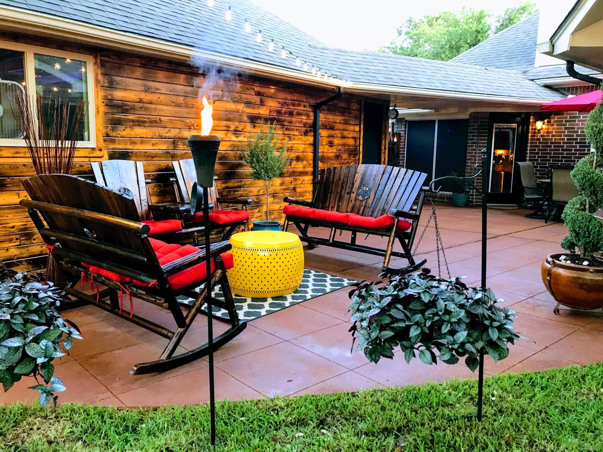 A vibrant scored concrete patio stained with a mix of Milano Red, Driftwood, and Pumpkin Antiquing concrete stains, complemented by eye-catching red wooden patio chairs, a vivid yellow table, torches, and string lights for a lively outdoor atmosphere.