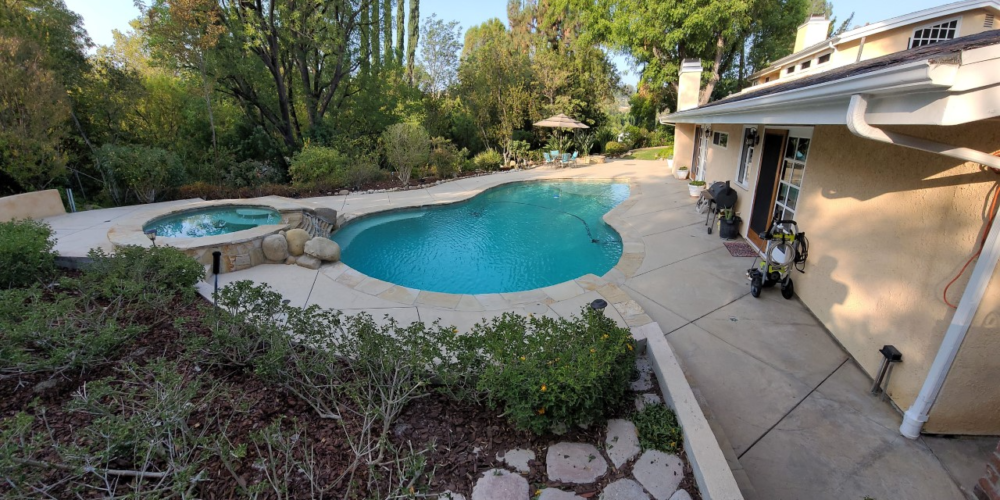 A before photo showing a wide expanse of faded and colorless stamped concrete around a pool, fire pit, and extending to other areas of the backyard