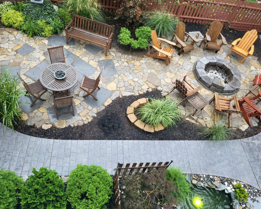 An aerial view of a spacious patio deck featuring a firepit area with chairs, a round outdoor table area, and a paver walkway stained with light charcoal concrete. The patio deck floor is made of natural stones in various sizes, stained in Tweed, Charcoal, and Silver Gray concrete stains, creating a serene and inviting atmosphere.