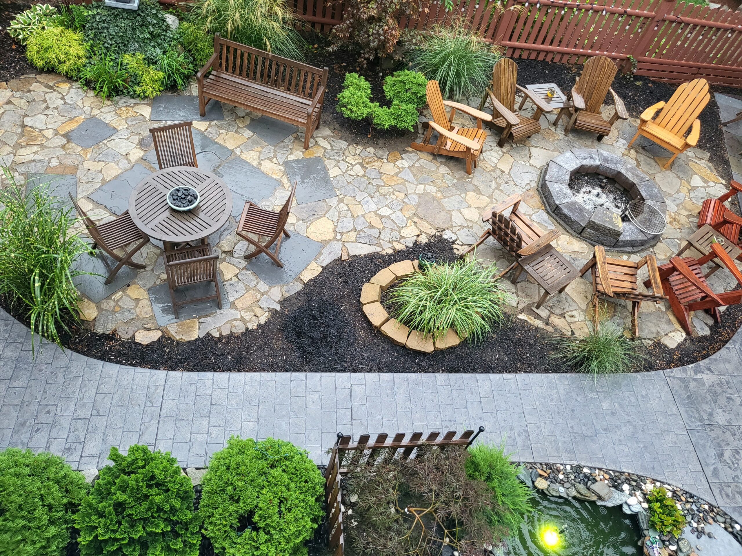 An aerial view of a spacious patio deck featuring a firepit area with chairs, a round outdoor table area, and a paver walkway stained with light charcoal concrete. The patio deck floor is made of natural stones in various sizes, stained in Tweed, Charcoal, and Silver Gray concrete stains, creating a serene and inviting atmosphere.