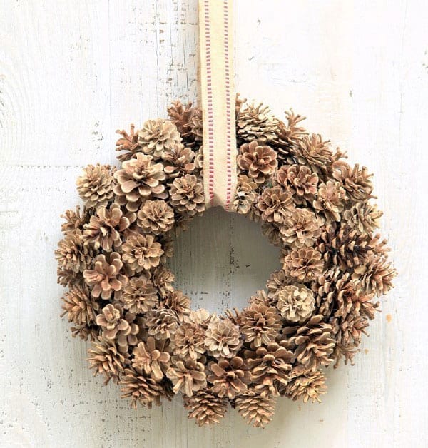 A pinecone wreath hangs by a burlap ribbon against a white wall.