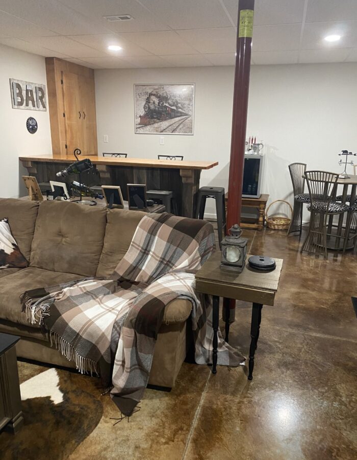 Final look of a transformed basement into a rustic family/rec room with desert amber and coffee brown acid stain. The room features a live edge bar area, sliding barn door, and a reclaimed wood feature wall.