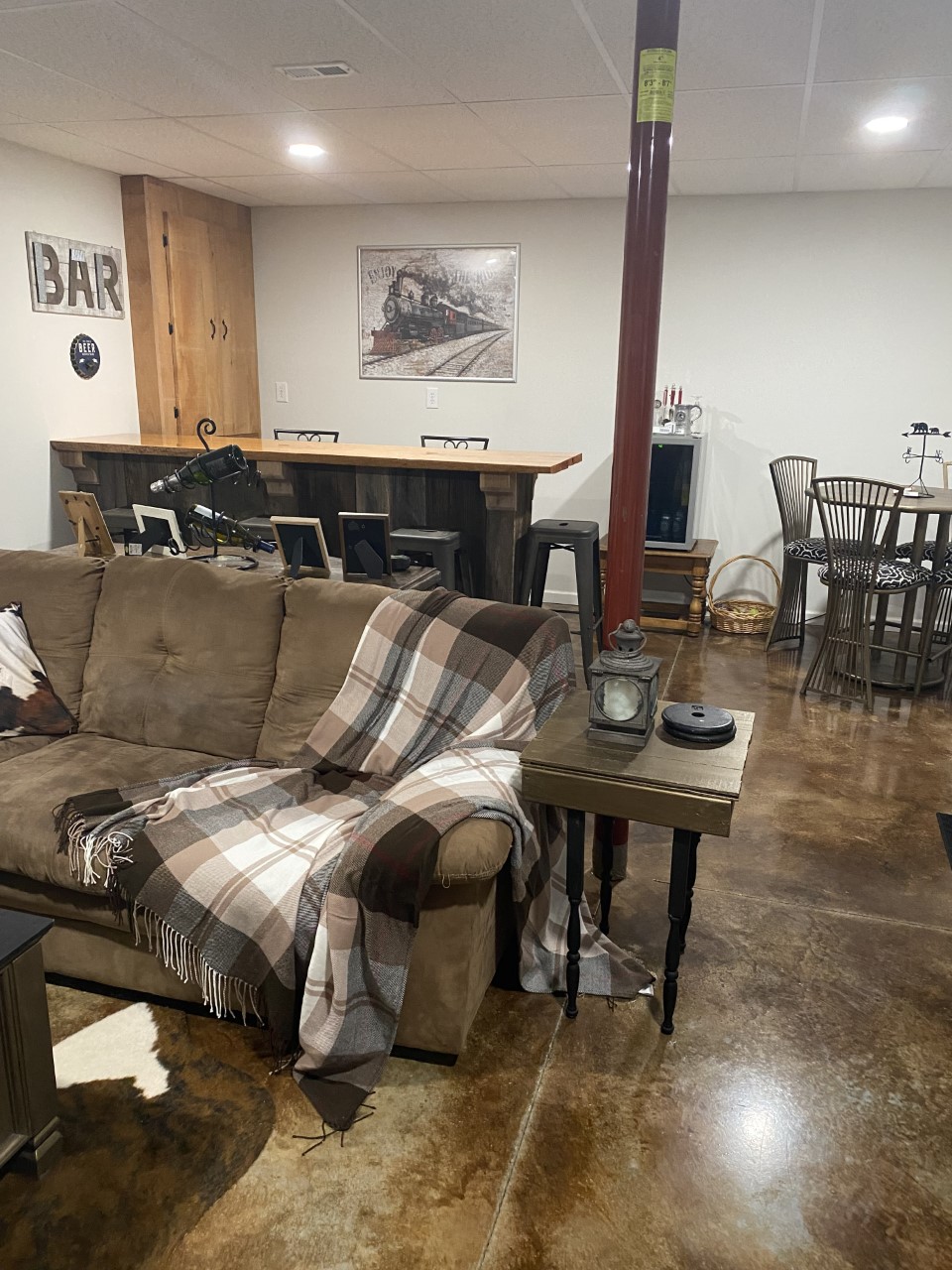 Final look of a transformed basement into a rustic family/rec room with desert amber and coffee brown acid stain. The room features a live edge bar area, sliding barn door, and a reclaimed wood feature wall.