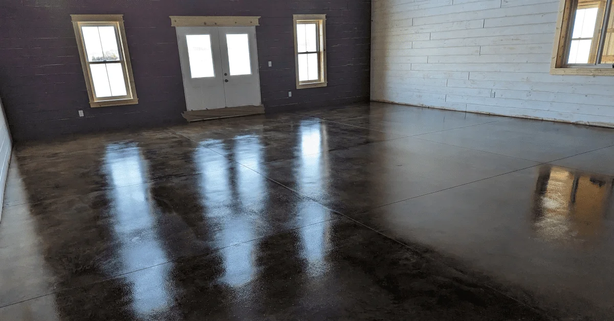 A room featuring a concrete floor with a black acid stain finish, resulting in a dark, mottled appearance with natural light creating soft reflections on the surface