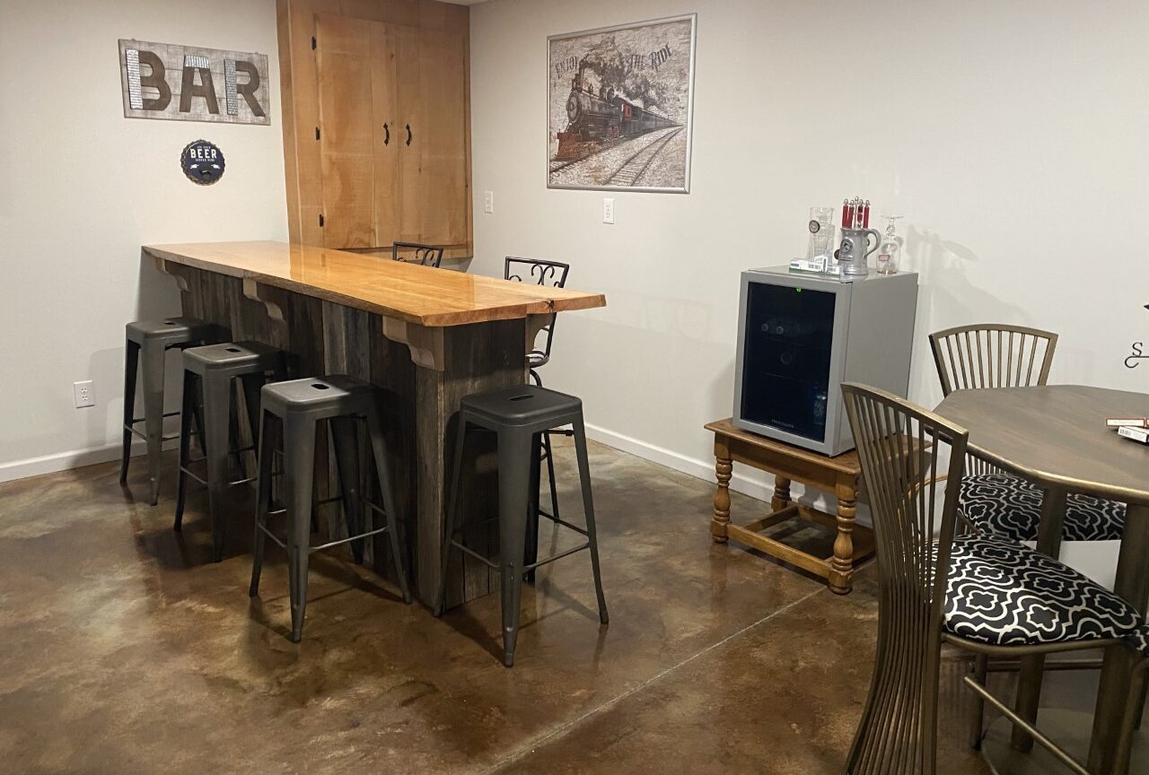 A basement bar with EverStain Coffee Brown and Desert Amber acid-stained concrete floors, featuring a smooth, leather-like finish. The space includes a wooden bar, metal stools, and a small refrigerator.