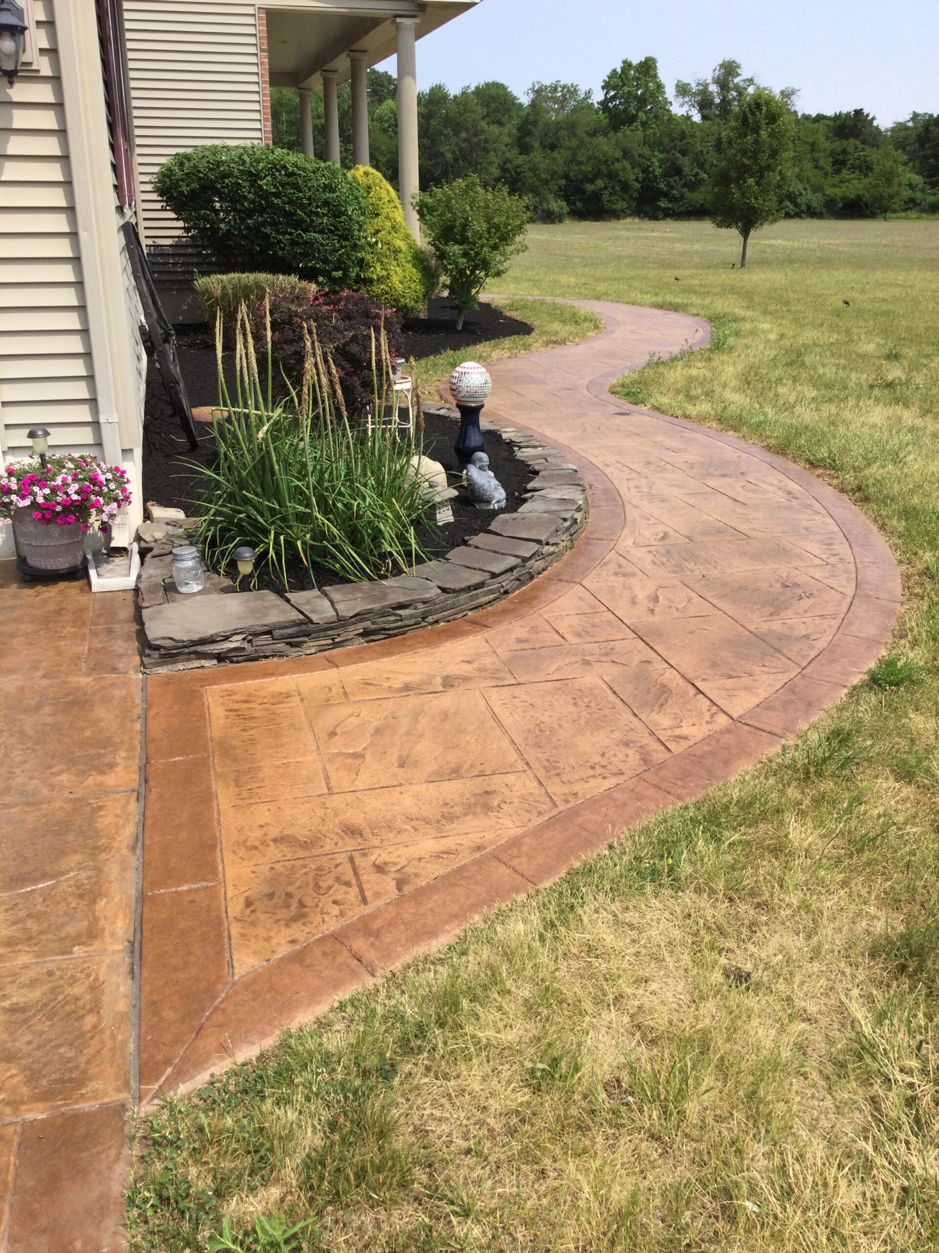 Curved stamped concrete walkway enhanced with cafe royale and driftwood antiquing stains, bordered by manicured garden beds and lawn