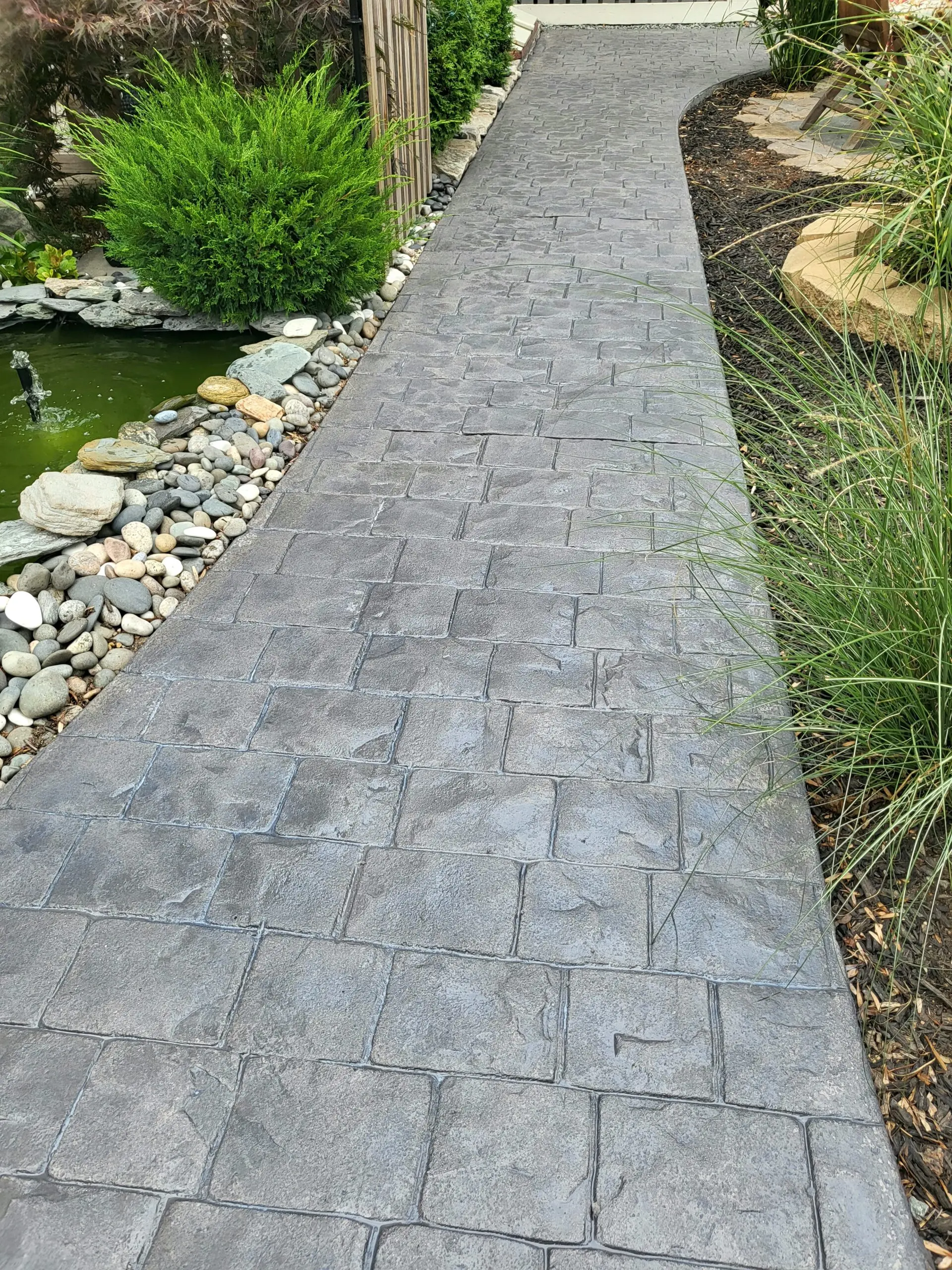 A narrow stamped concrete walkway stained gray runs alongside a tranquil garden pond