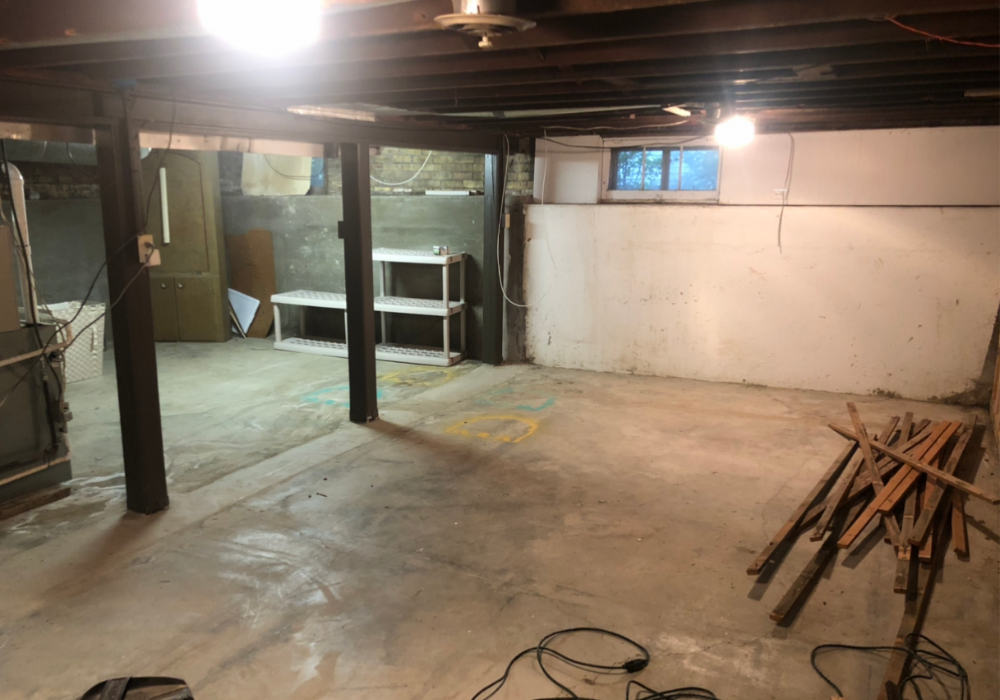 An unfinished basement with exposed beams, a stained concrete floor, and scattered construction materials