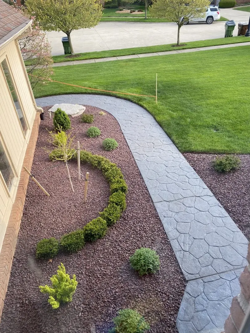 Overhead view of a curved stamped concrete walkway stained in eagle gray, surrounded by ornamental plants and pebbles