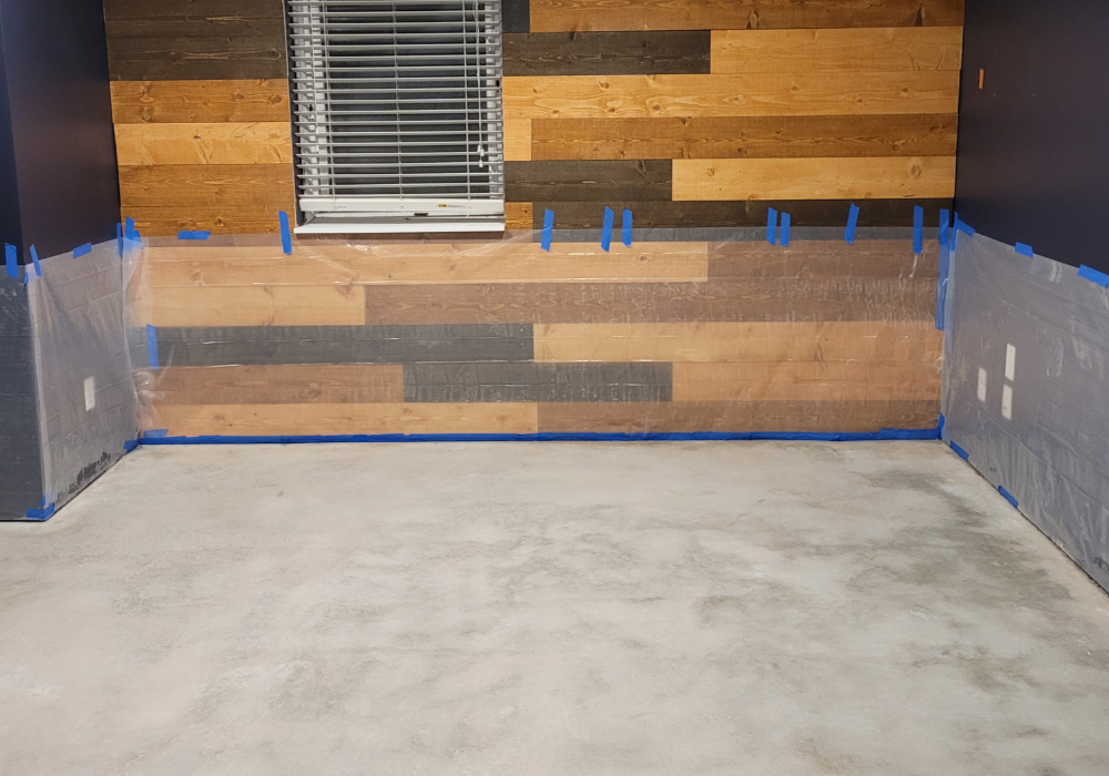 A prepared basement floor with carpet removed, cleaned concrete, and plastic sheeting taped to the walls before applying black acid stain