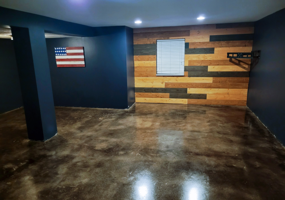 A modern basement man cave with a black acid-stained concrete floor, dark blue walls, and a rustic wood accent wall