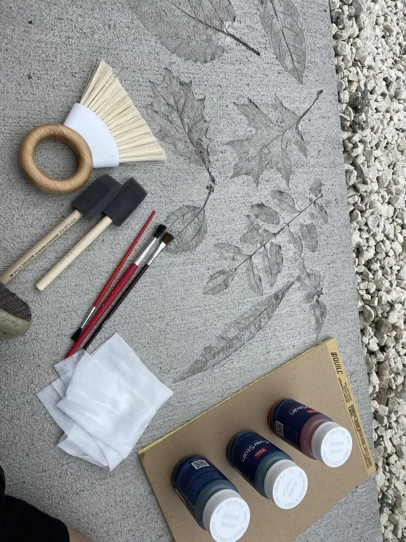 Close-up of tools including brushes, foam applicators, and EverStain acid stain bottles, with leaf stencils on concrete in the background.