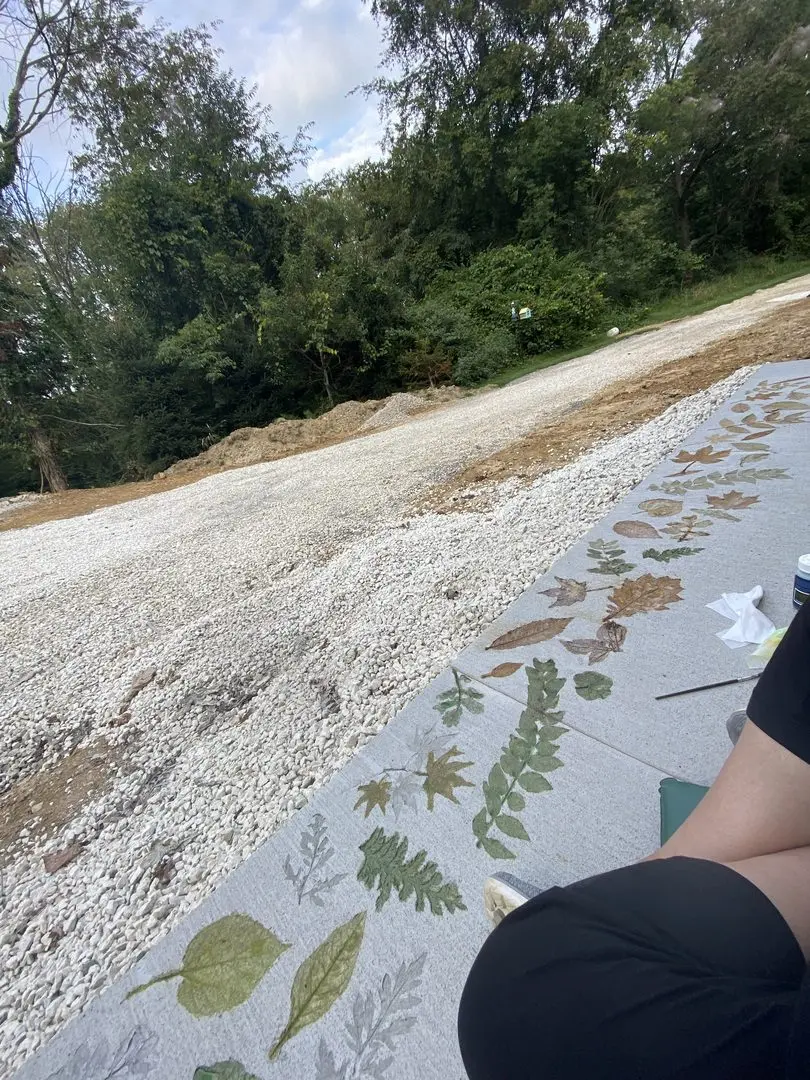 Concrete walkway with multiple leaf stencils in green and brown tones created with EverStain acid stains, extending along a gravel driveway in a natural setting.