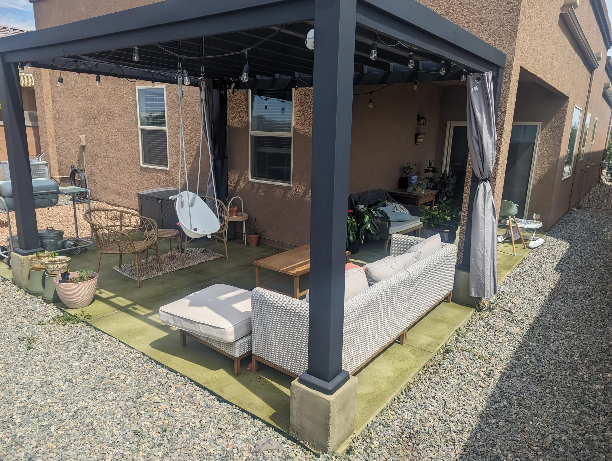 A modern covered patio with concrete flooring stained in ColorWave Olive, featuring outdoor furniture, potted plants, and a swing chair, blending natural tones with the surrounding design.