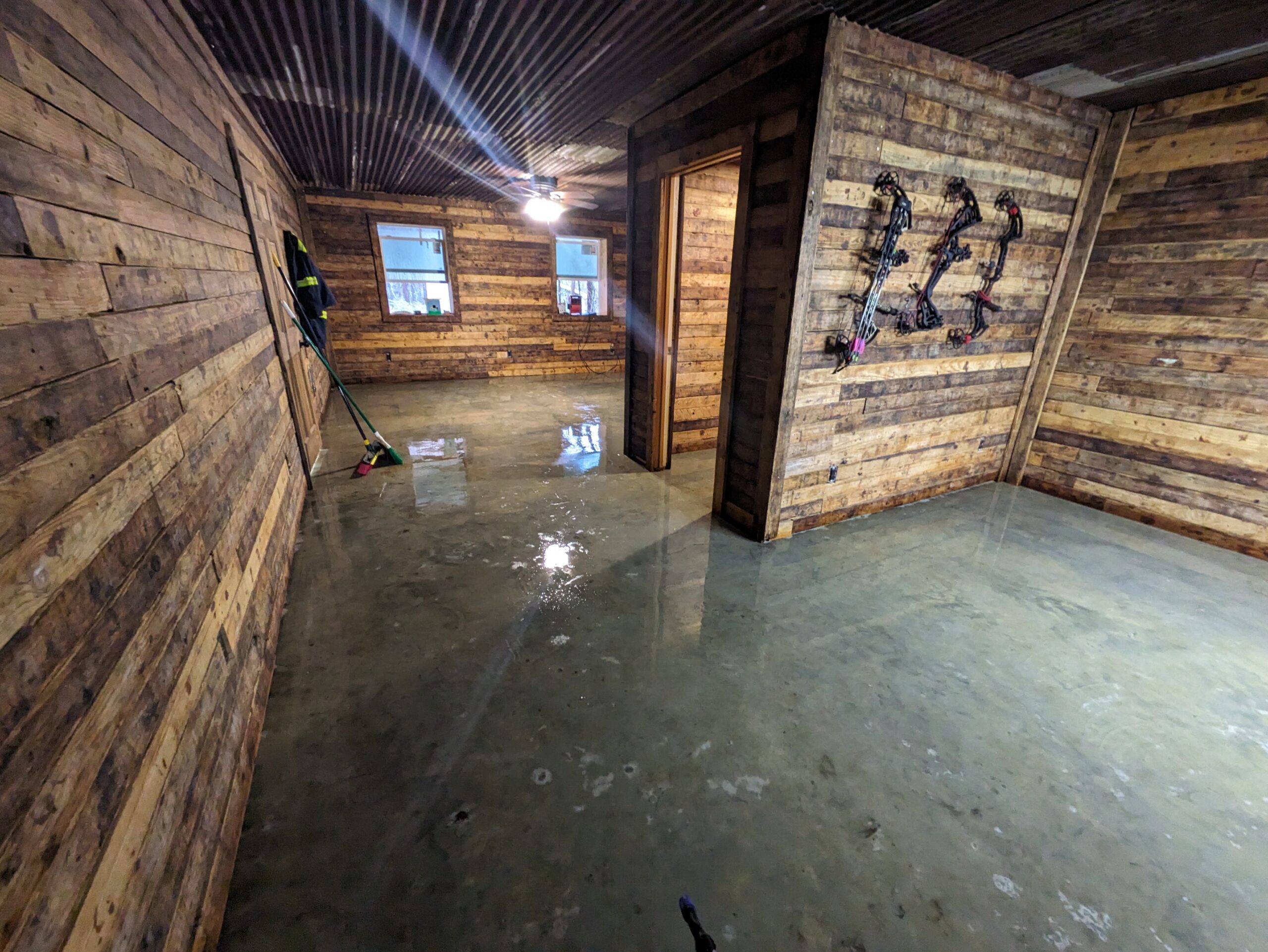 A room with rustic wooden walls and a plain, smooth concrete floor before staining, with tools and hunting bows visible on the walls.