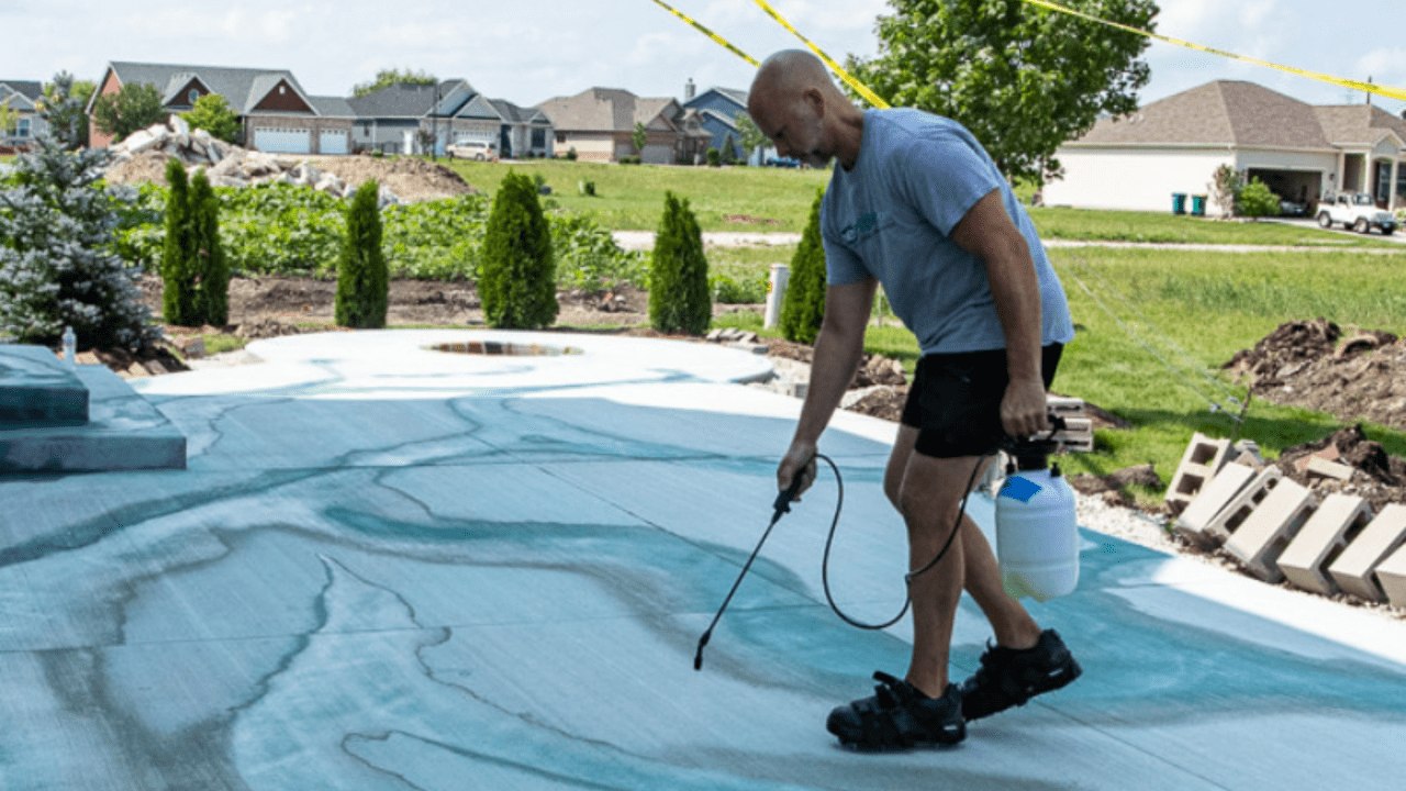 A person spraying an acid stain solution on a concrete surface outdoors, with swirling blue-green patterns forming on the ground.