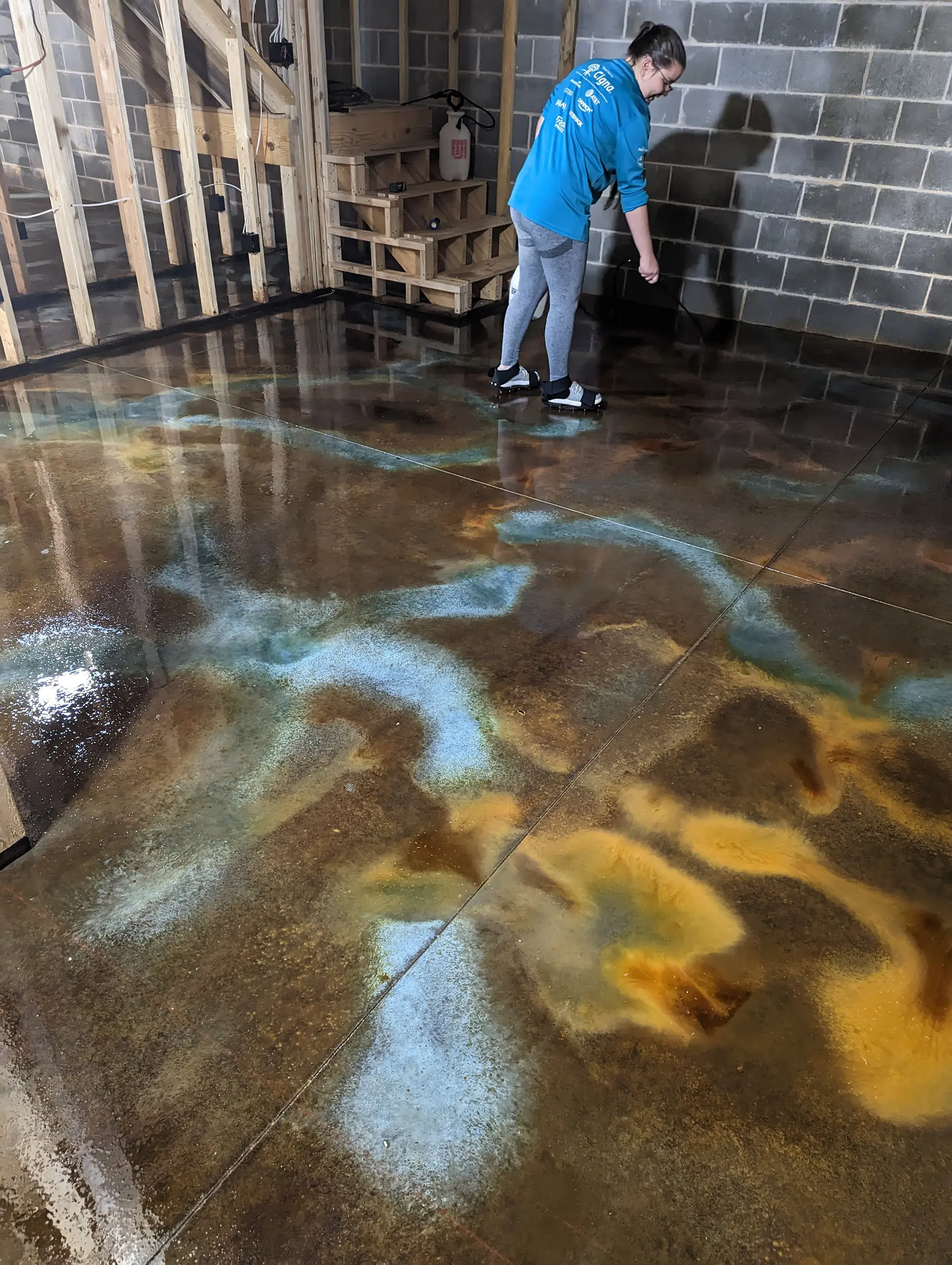 Woman applying three different acid stains - Coffee Brown, Malayan Buff, and Seagrass - onto the concrete basement floor using a pump sprayer