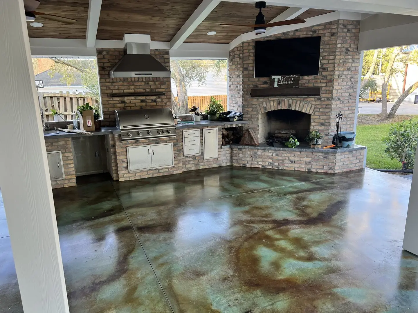An image of a spacious outdoor porch featuring acid stained concrete floors in shades of Malayan Buff and Seagrass. The scene displays an elegant outdoor kitchen setup in the background