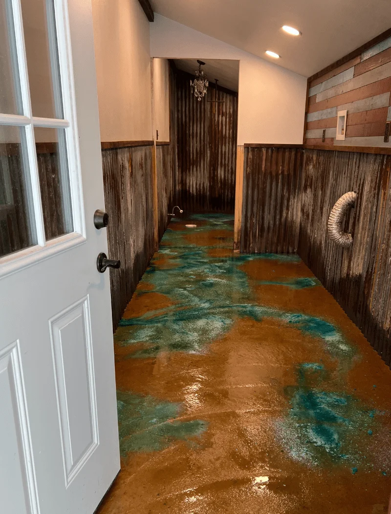 An eclectic bathroom with a vibrant, acid-stained concrete floor in hues of azure blue and earthy tones, blending a rustic wooden wall finish with a splash of modern color